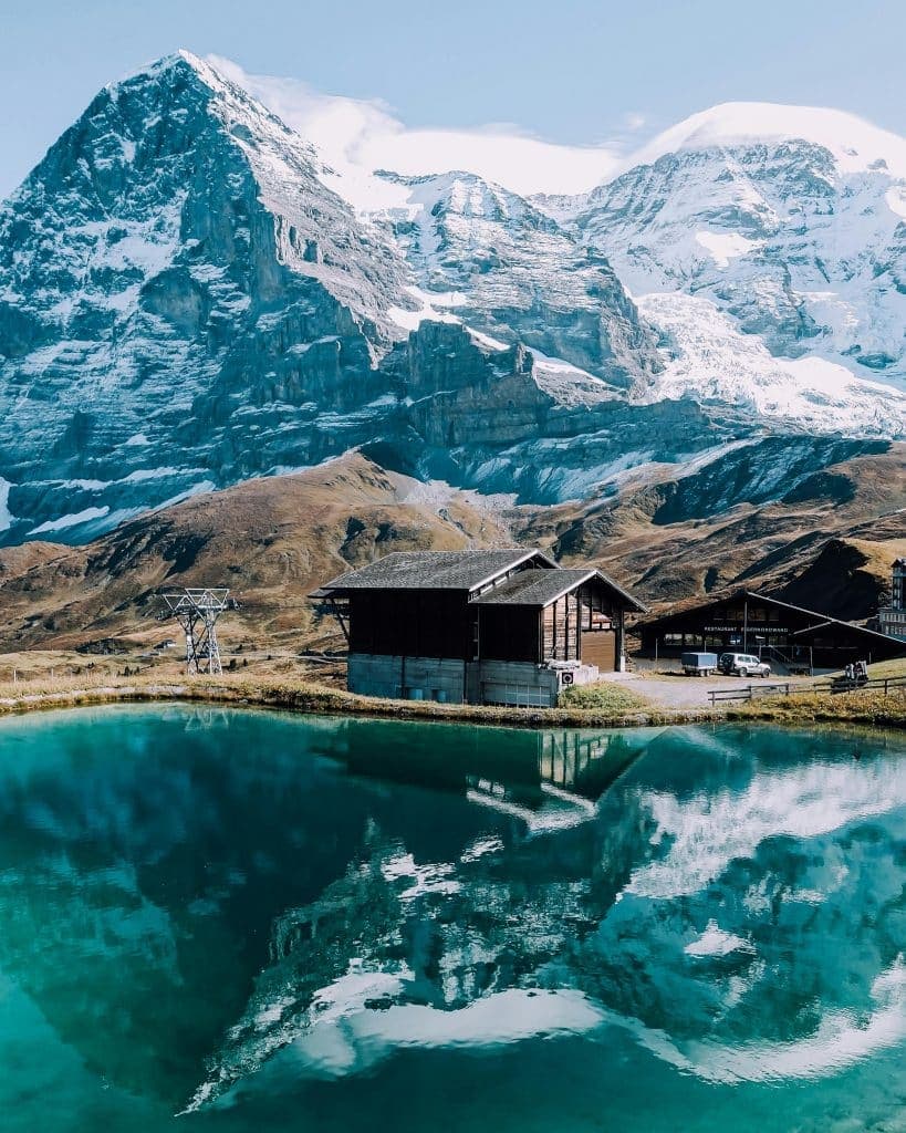 Snow-capped mountains reflect in a tranquil turquoise lake, flanked by rustic wooden cabins on grassy terrain. This Swiss scene exudes serenity, capturing the natural beauty of the rugged landscape under a clear blue sky.