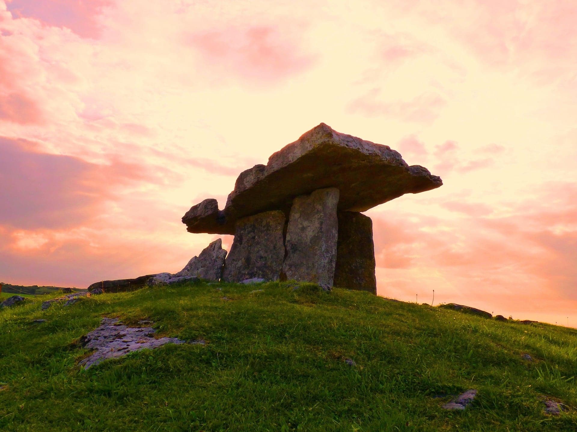 A large ancient stone dolmen stands on a grassy hill under a dramatic sunset sky, with pink and orange clouds illuminating the scene—a perfect highlight for Steelers in Dublin Travel Packages.