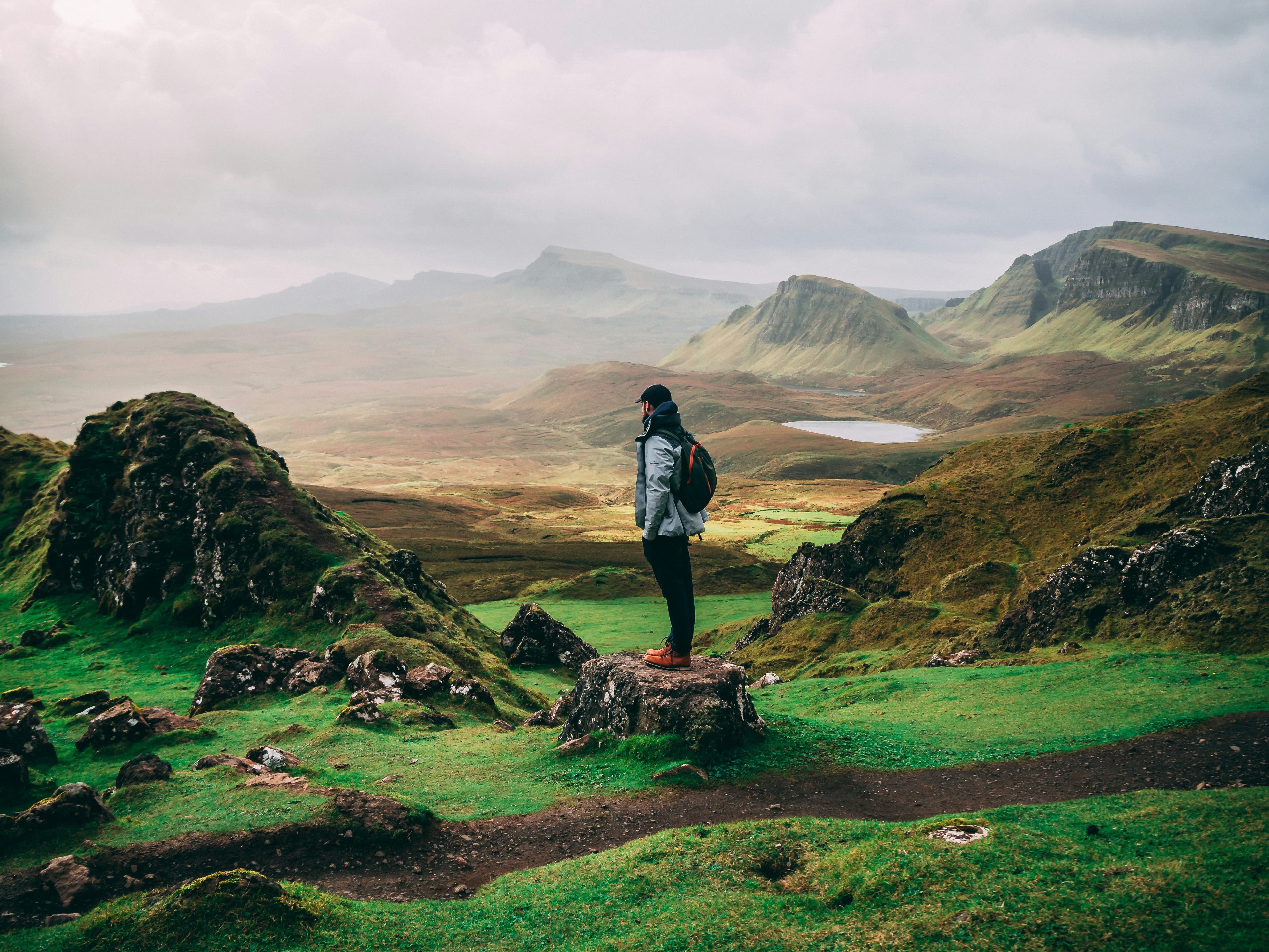 A person wearing a jacket and backpack stands on a rock, overlooking a lush green valley with rolling hills and distant mountains under a cloudy sky.