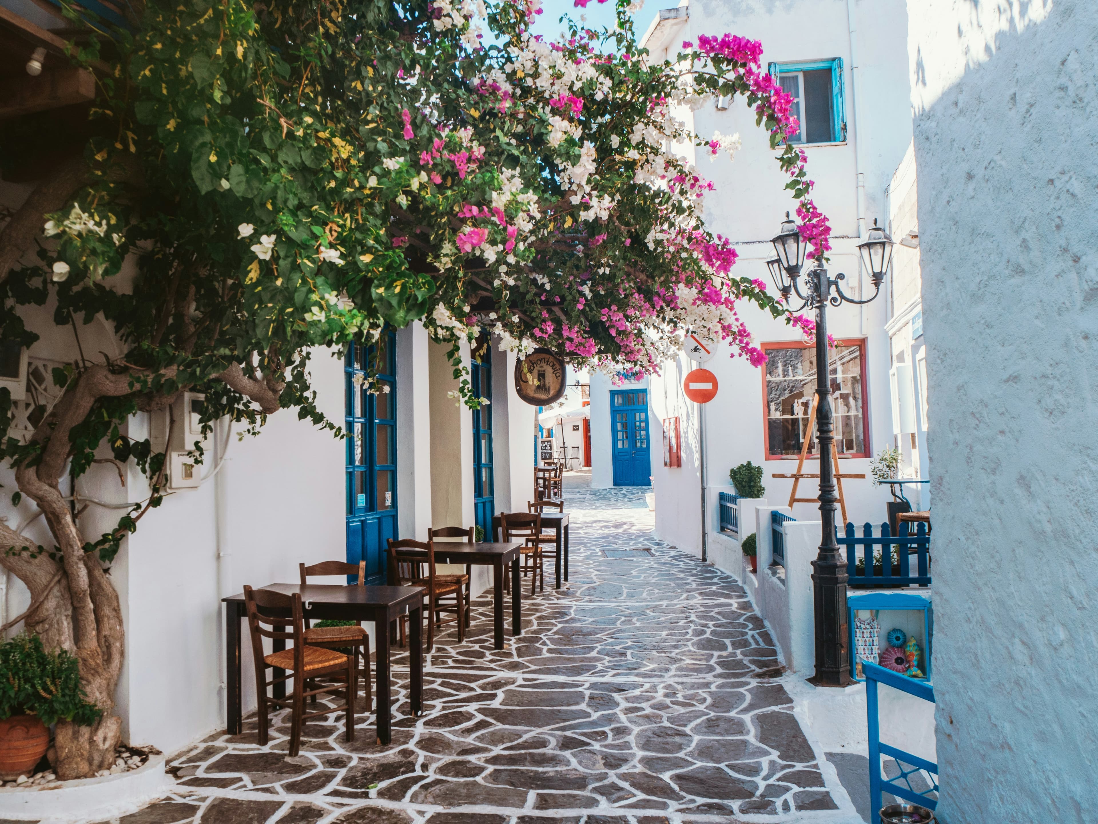 A narrow cobblestone street in a Greek village lined with whitewashed buildings, blue doors and windows, outdoor tables and chairs, and vibrant pink bougainvillea overhead—a scene as charming as how Ireland got the name the Emerald Isle.