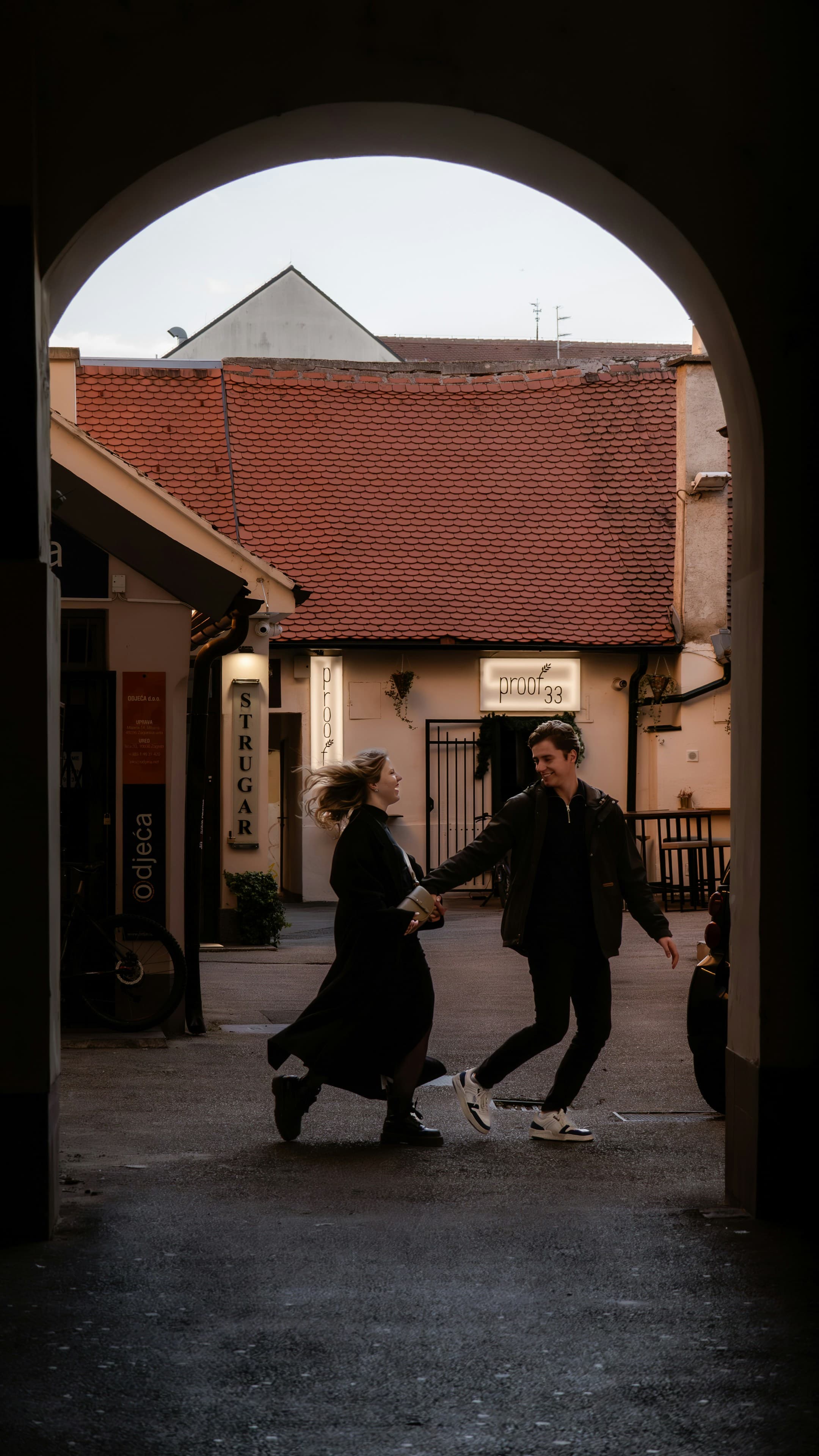 A couple holding hands runs playfully through an archway into a charming courtyard with red tiled roofs and small shops, lit by soft, natural daylight—capturing the romance found on Custom Croatia Tours.