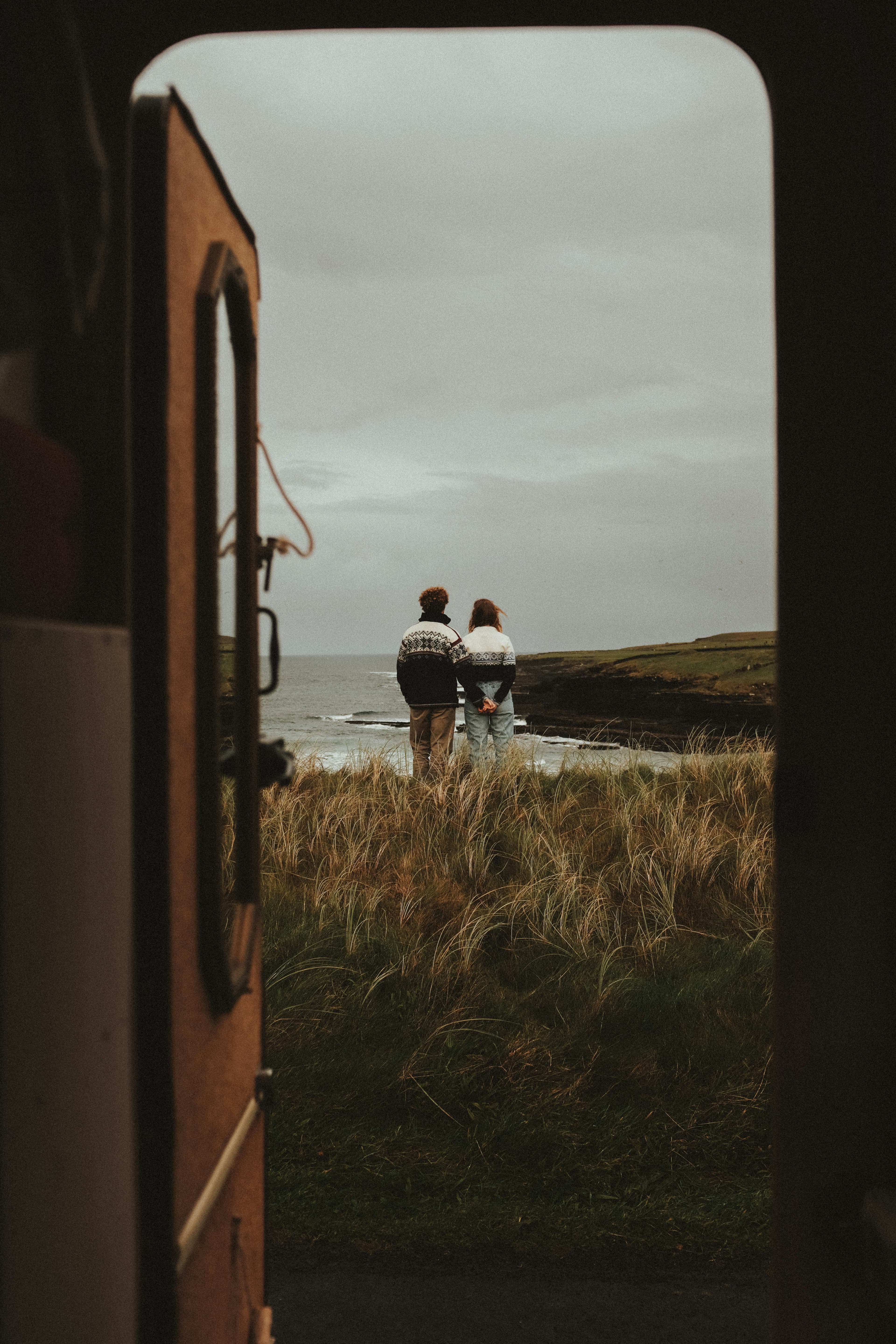 A couple stands close together on grassy terrain, facing the ocean as seen from the open door of a vehicle under a cloudy sky, capturing the romance of an Italy and Greece Honeymoon Itinerary.