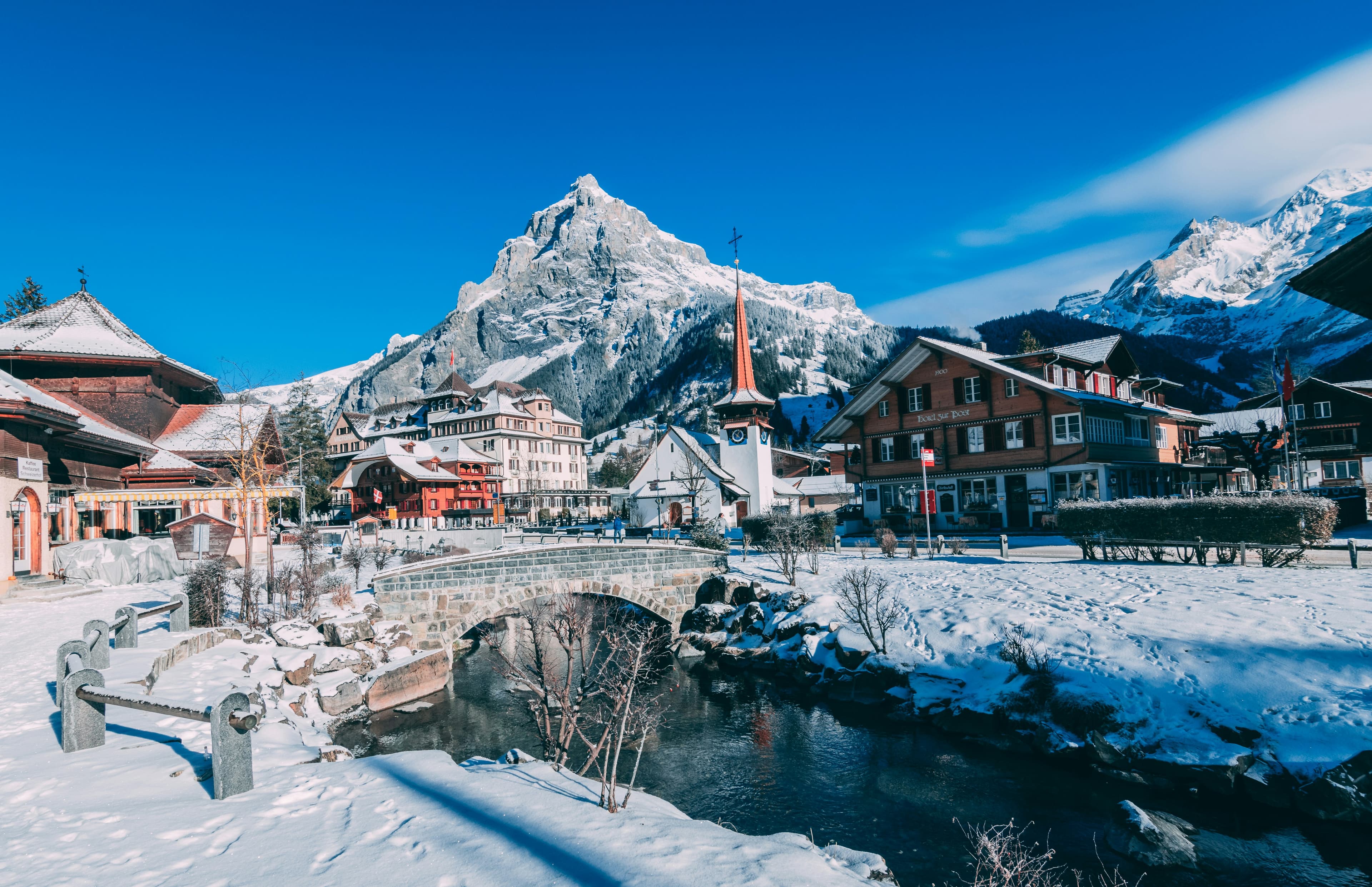 A picturesque snowy village with Alpine style houses, a church steeple, stone bridge, and stream, set against snow covered mountains—perfect for inspiring your Italy and Greece Honeymoon Itinerary under a clear blue sky.