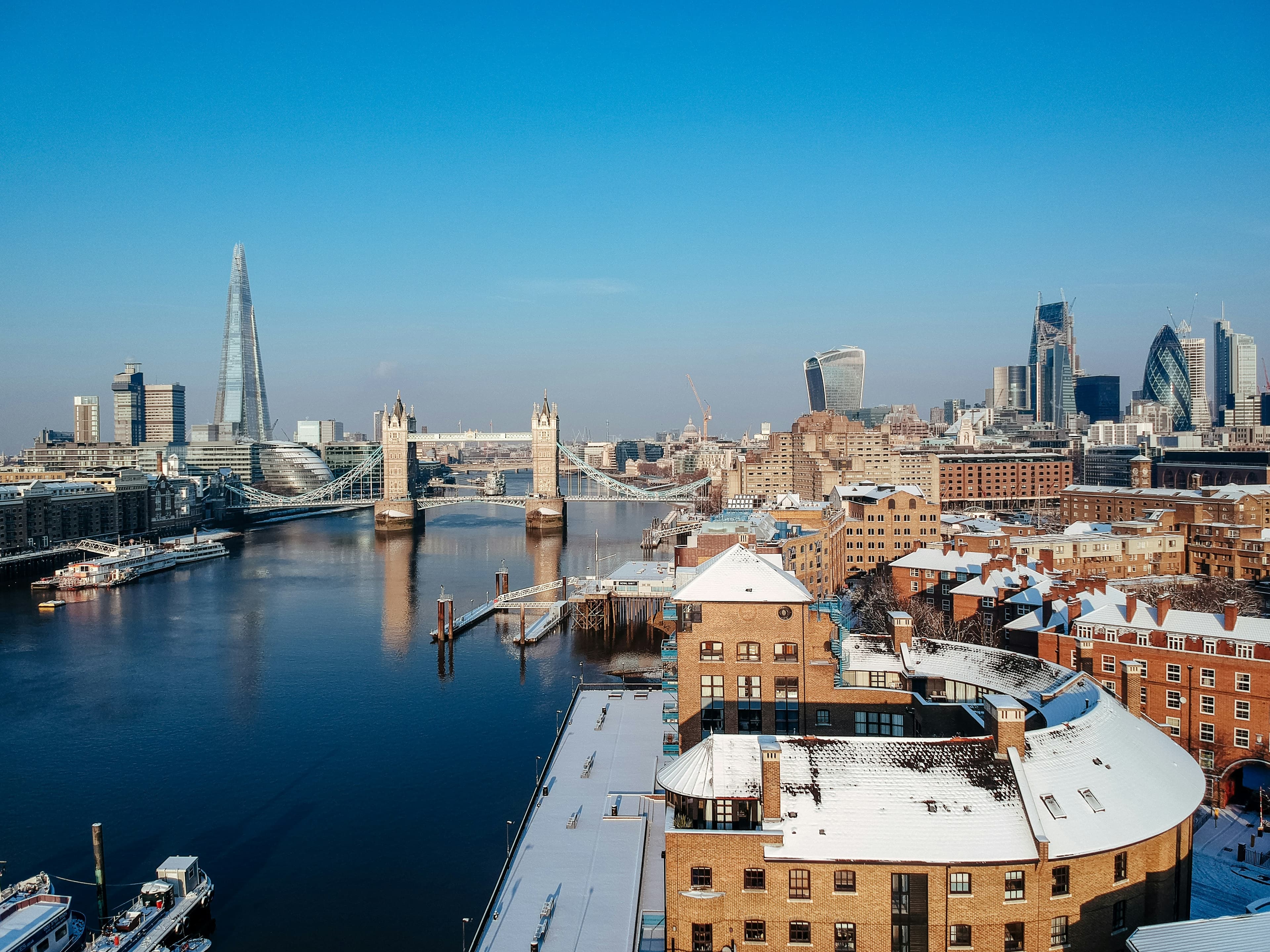 Aerial view of London featuring the River Thames, Tower Bridge, The Shard, and modern skyscrapers under a clear blue sky. Some rooftops along the river are dusted with snow—perfect inspiration for custom vacation packages.