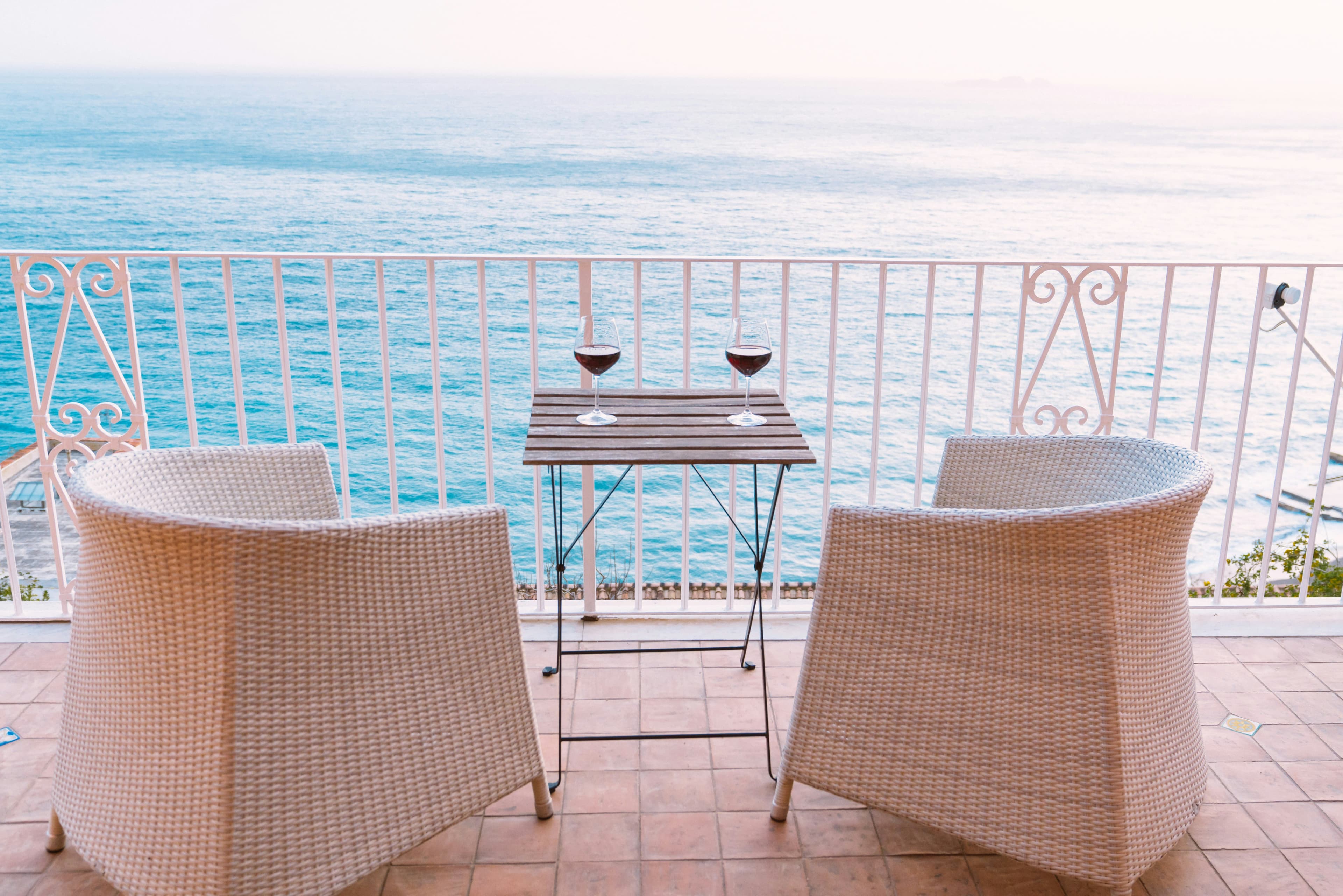 Two wicker chairs face a small wooden table with two glasses of red wine on a tiled balcony, overlooking the calm, blue ocean under a pale sky—perfect for reflecting on what is Victory Day in Croatia and its meaning.