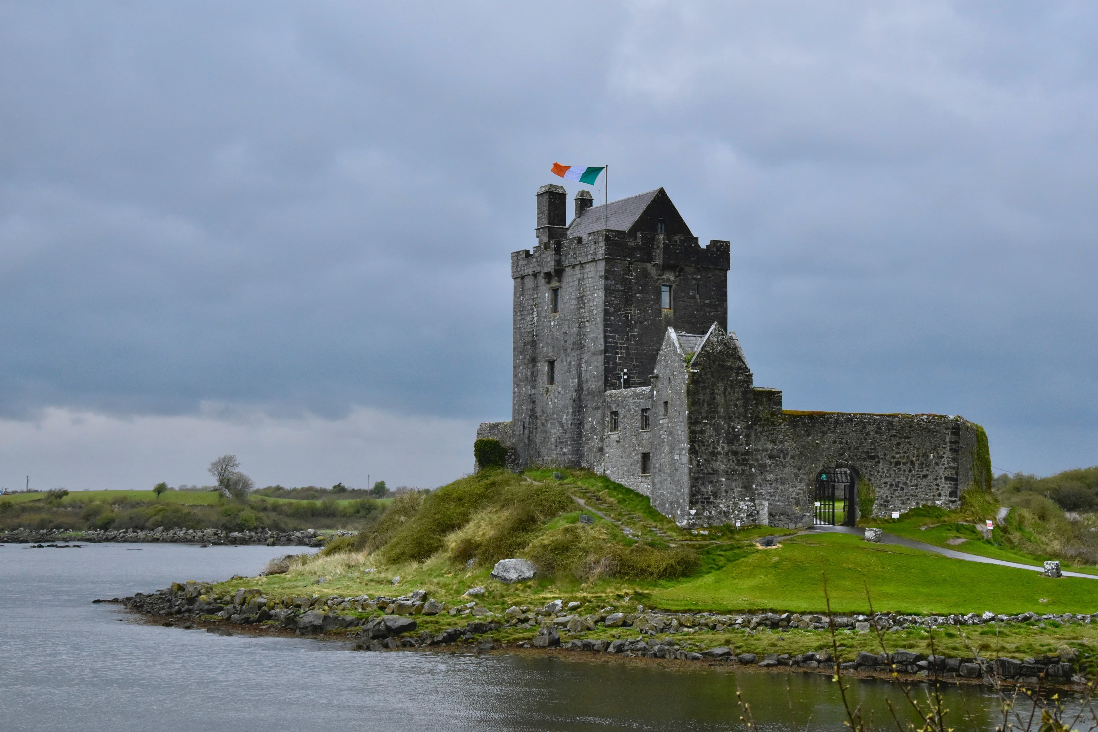 A stone castle with two flags atop sits on a grassy, rocky peninsula by the water under a cloudy sky, surrounded by green fields and distant trees—a perfect gaff, as they say in Irish slang.