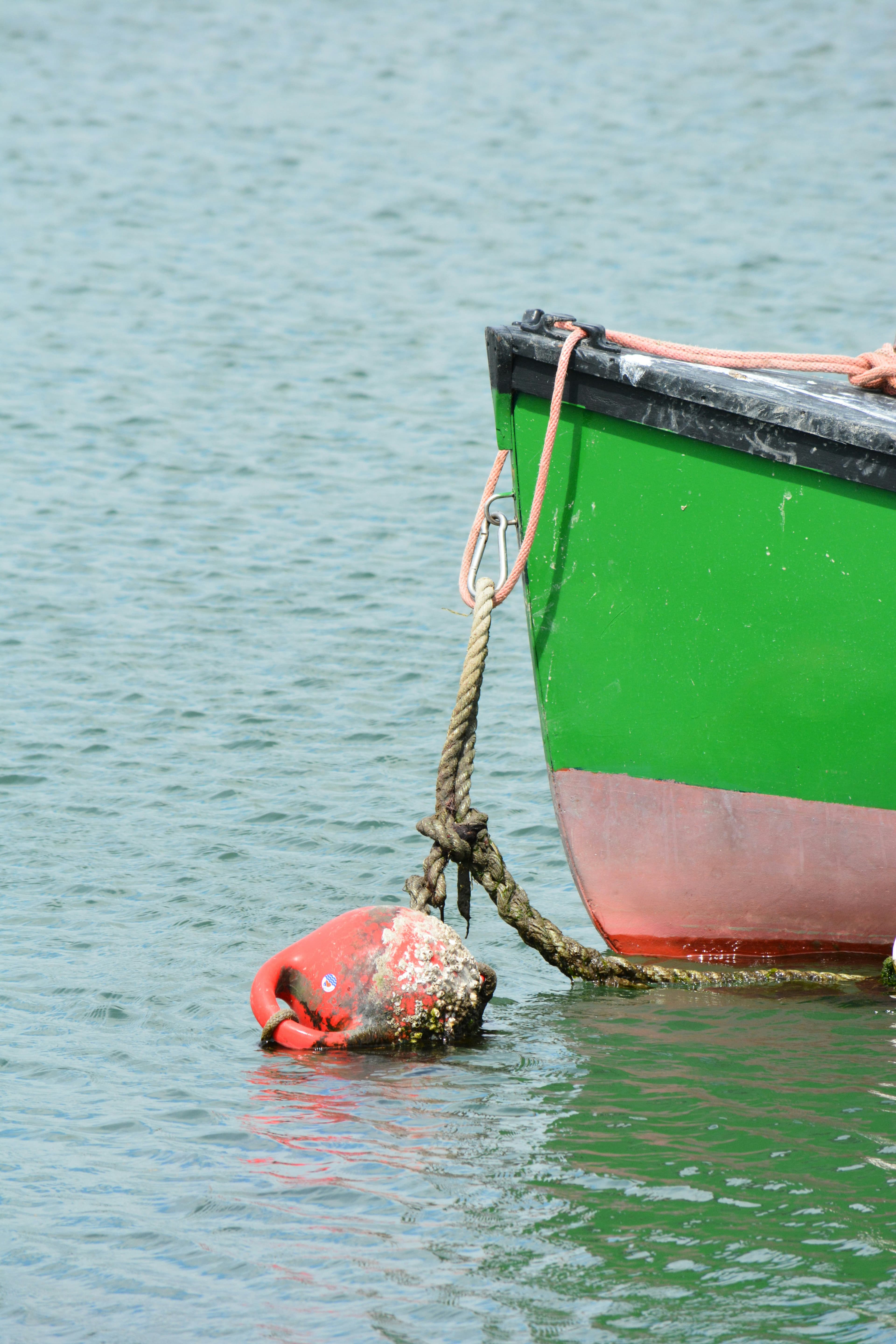 A green boat is tied to a weathered red buoy floating on calm water. The boat’s bow and rope are visible, with the water reflecting light ripples.
