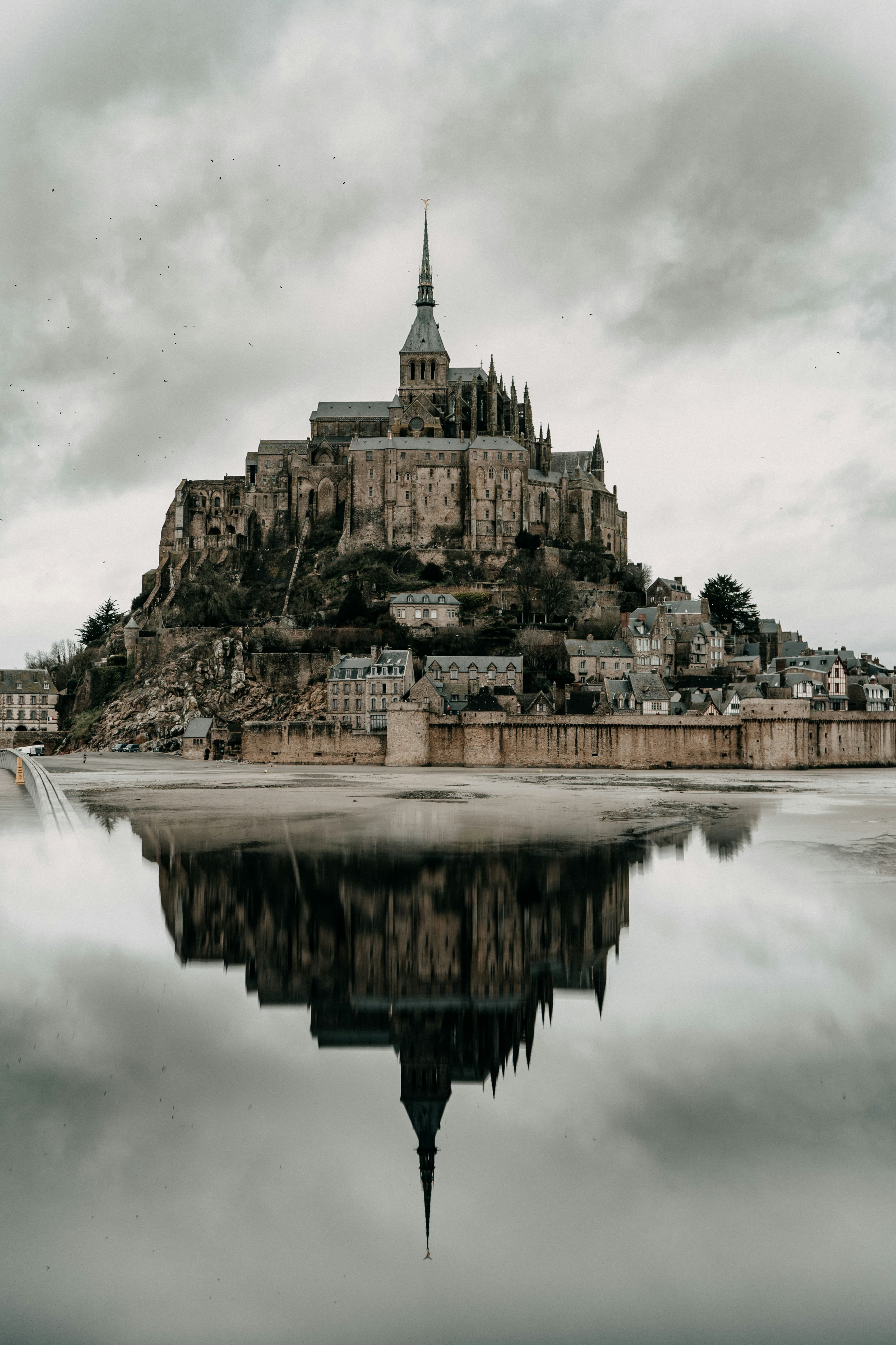 A medieval abbey sits atop a rocky hill, surrounded by small village buildings, with its reflection visible in calm water under a cloudy sky.