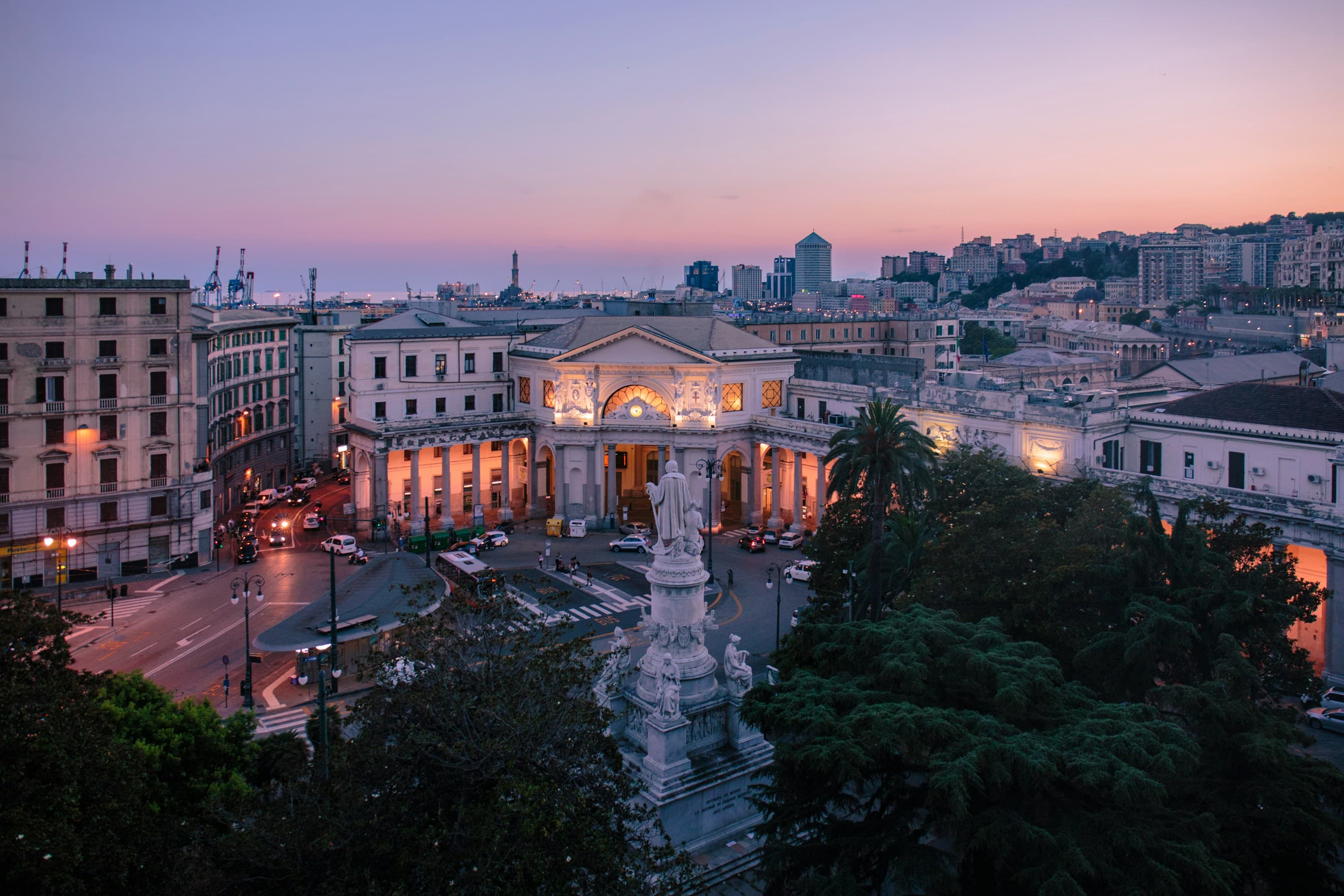 A city square at sunset with a grand, illuminated neoclassical building, statue in the foreground, historic buildings, trees, and a skyline under a pink and purple sky—perfect for couples exploring Best Europe Honeymoon Packages 2026.