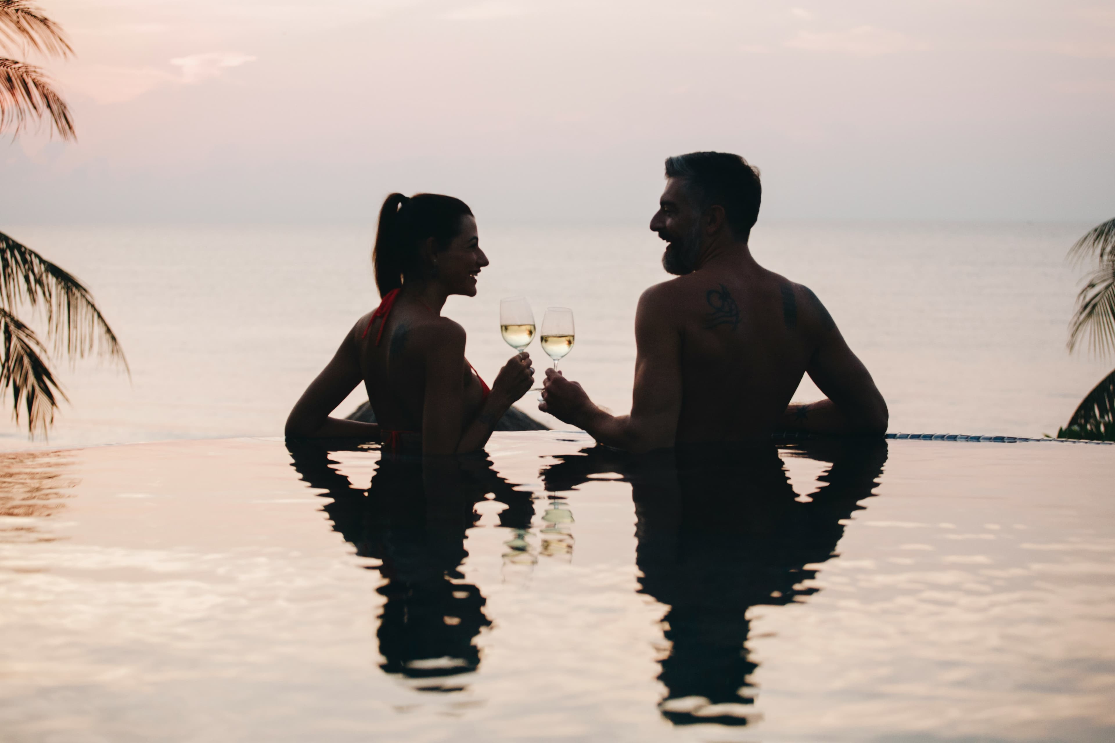 A couple in swimwear enjoys glasses of white wine while relaxing in an infinity pool at sunset, with the ocean and palm trees in the background—capturing the essence of Top Honeymoon Destinations 2024.