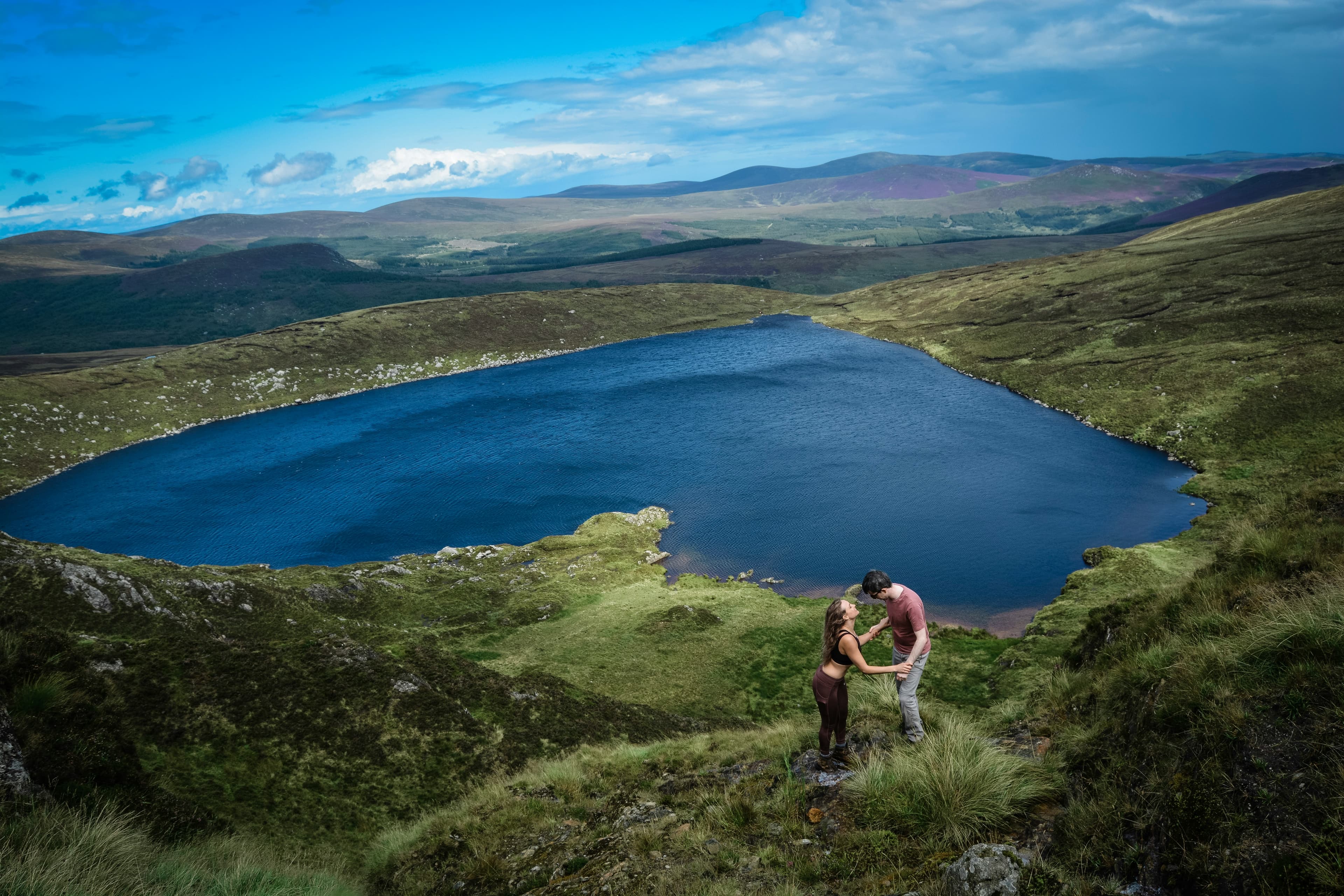 A couple stands holding hands on a grassy hillside, overlooking a heart shaped blue lake—a perfect spot for a romantic gaff, as the quintessential Irish slang would have it—surrounded by rolling hills under a bright sky with scattered clouds.