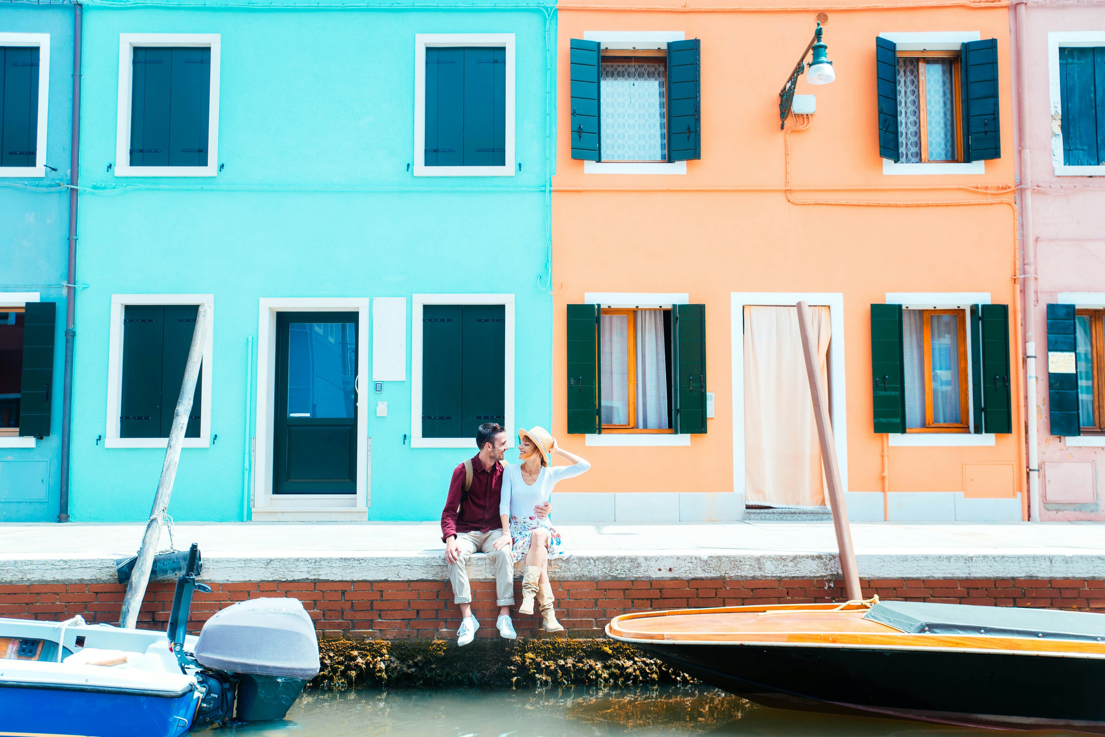 A couple enjoys their honeymoon on a canal edge, in front of colorful turquoise and orange houses with green shutters, next to docked boats on a sunny day—capturing the romance of Ireland or Scotland abroad.