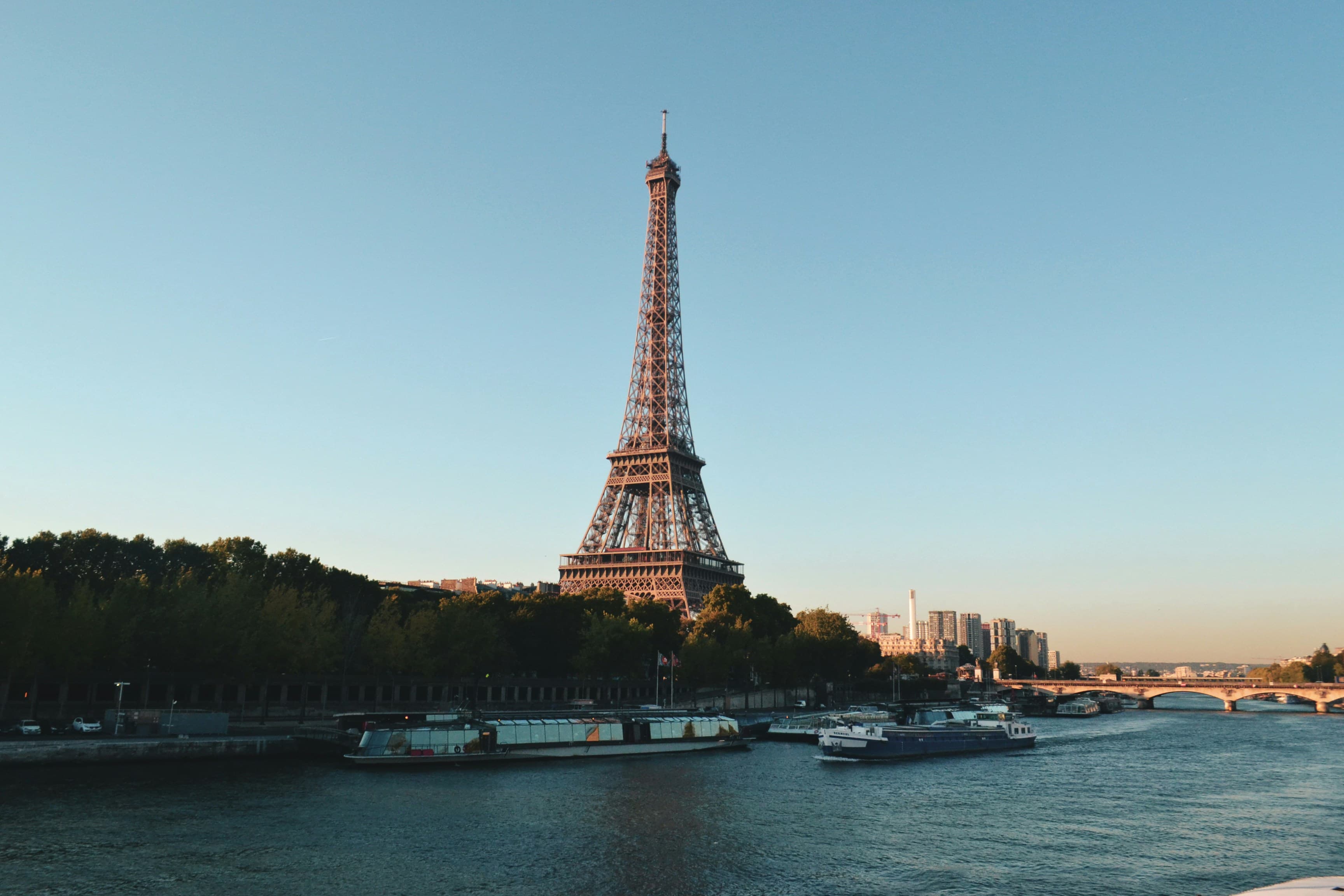 The Eiffel Tower stands tall by the Seine River in Paris, surrounded by trees and city buildings, with boats on the water under a clear blue sky at sunset.
