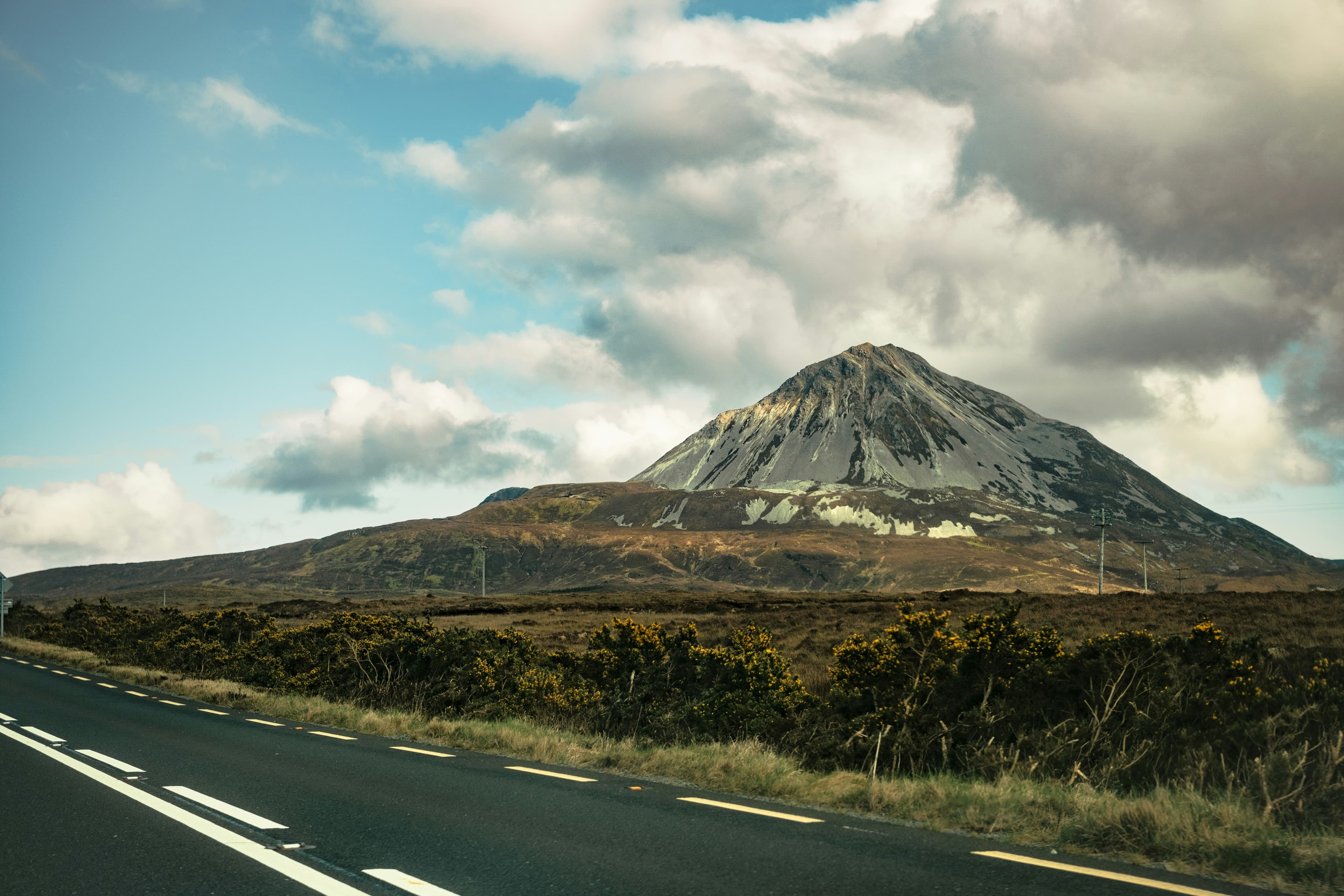 A two lane road curves alongside grassy fields and bushes, leading toward a large, rocky, and partly sunlit mountain beneath a cloudy blue sky.