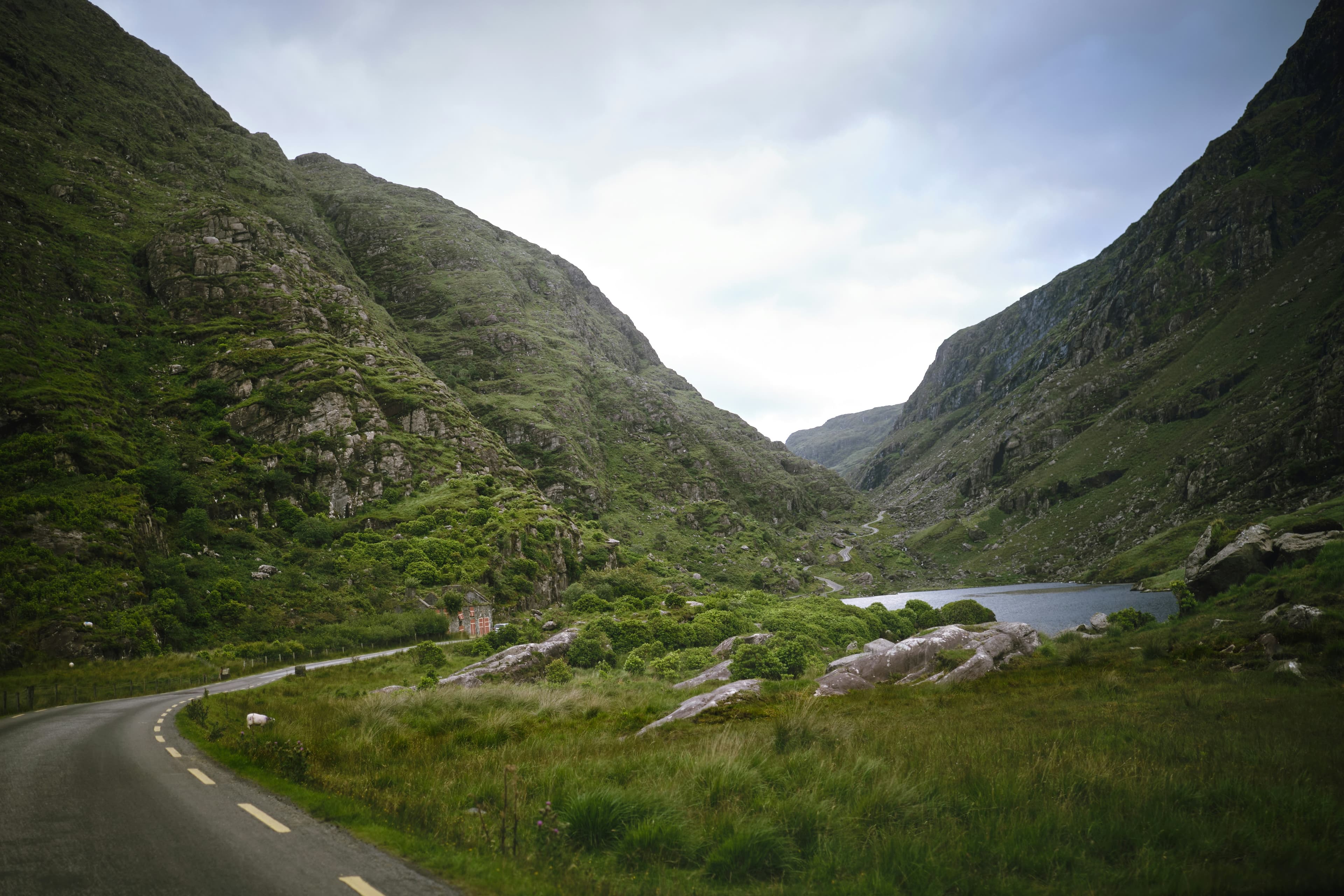A winding road curves through a lush green valley with rocky hillsides, a small lake, and cloudy sky in the background. Dense vegetation covers much of the landscape.