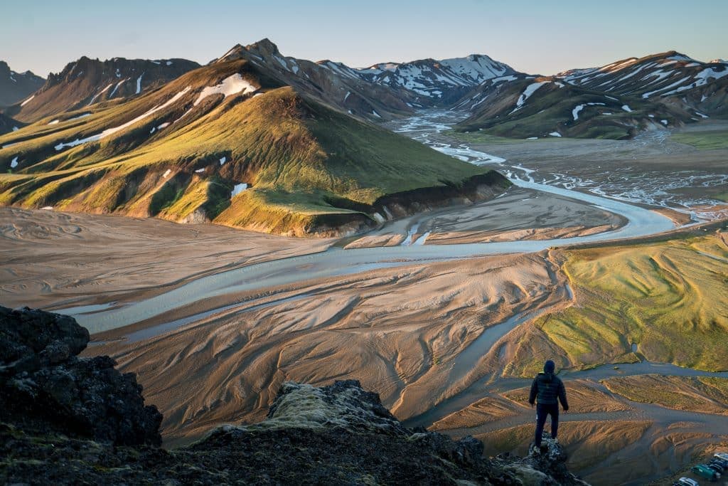 A person stands on a rocky outcrop, overlooking a vast landscape of meandering rivers and green mountains under a clear sky. Sparse patches of snow dot the scene, highlighting the rugged terrain—an emblem of Iceland's natural wonders.