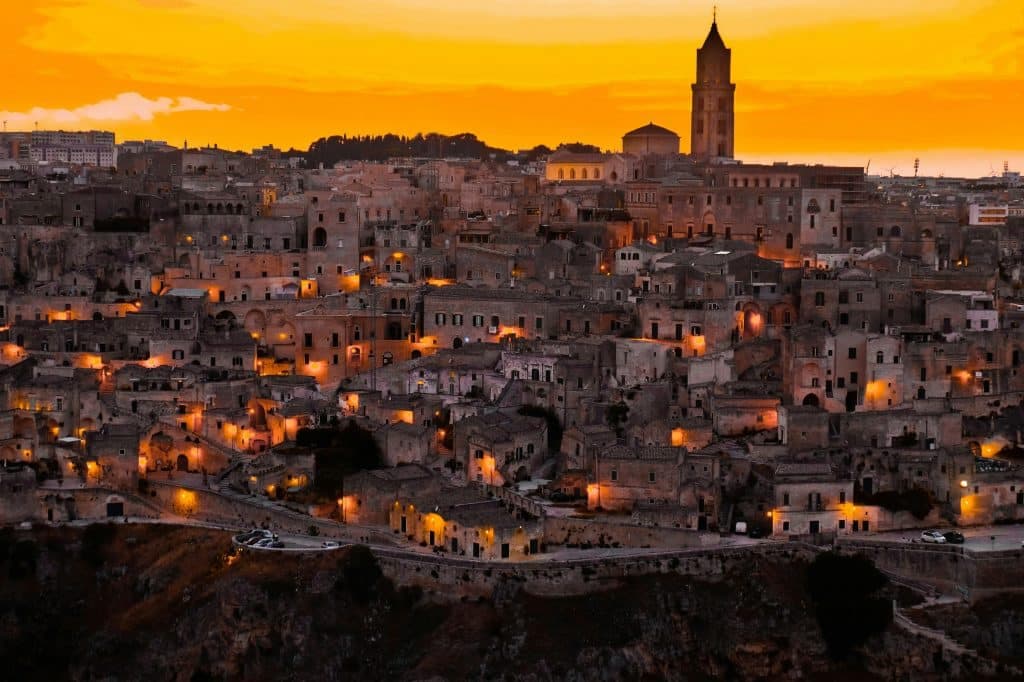 A stunning view of Matera at sunset, with the old city's stone buildings illuminated by warm, golden lights. A church tower rises prominently against the vibrant orange sky, creating a picturesque scene of this historic Italian city.