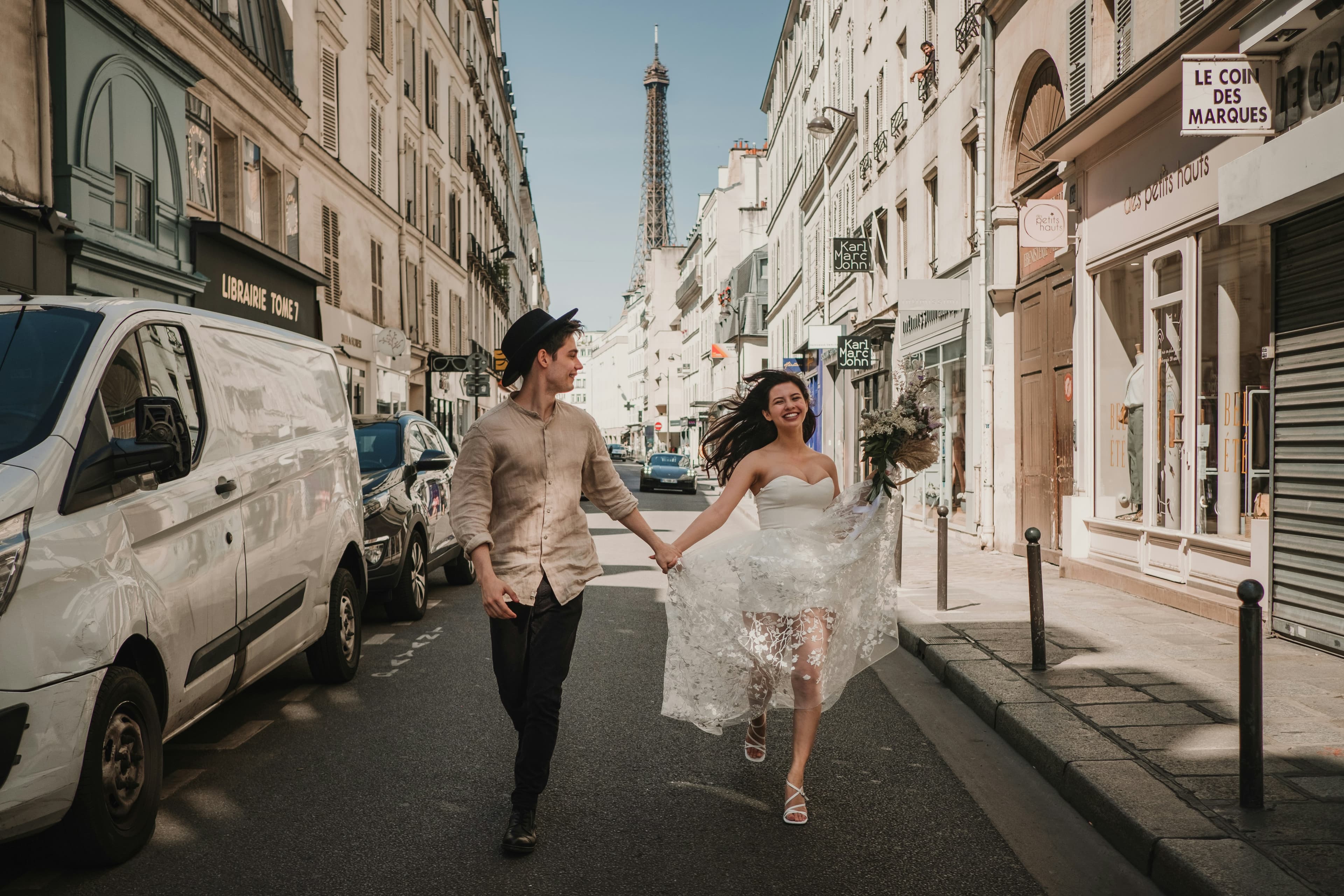 A joyful couple, with the Eiffel Tower in the background, runs hand in hand down a Paris street; the woman wears a white bridal dress and the man is dressed casually with a hat.